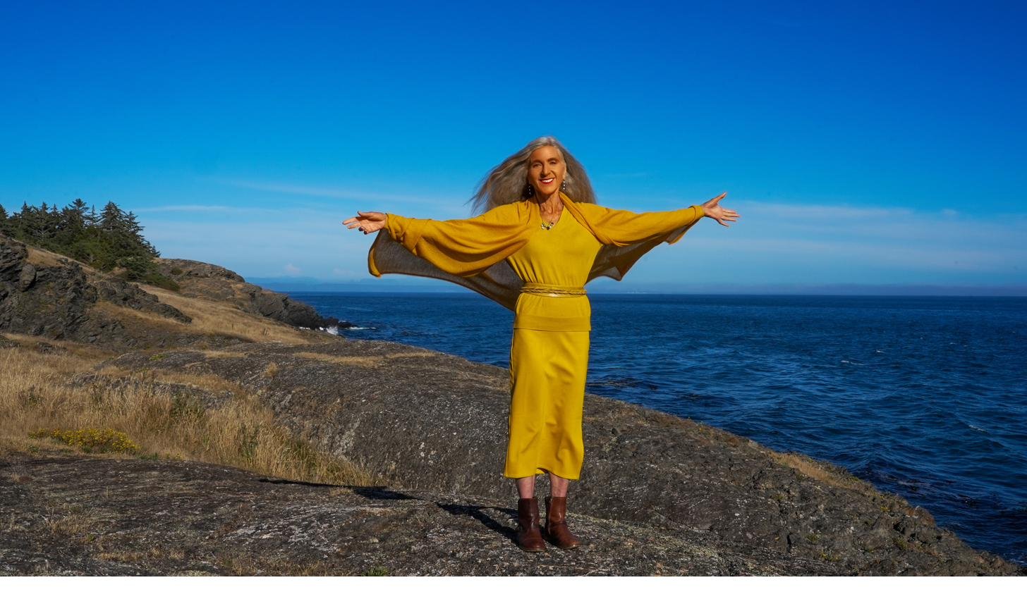 Iana Lahi standing with arms open in white linen by the sea